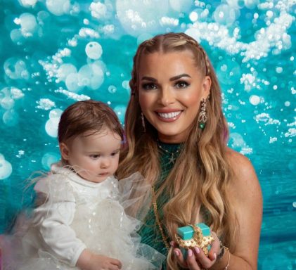 Vrouw met lang, blond haar en een kindje in een witte jurk, beide met kleurrijke decoratie.